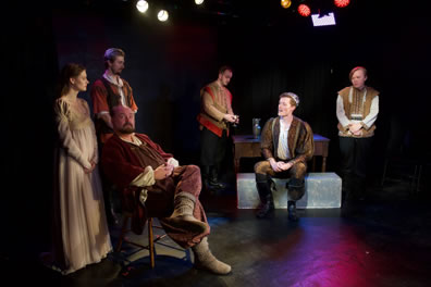 Prince Henry sits on a chair with legs crossed, looking up and away disinterestedly from King Philip sitting further back stage on a bench. Standing behind King Henry are Alais in simple tan gown and Richard; pouring a drink at the table behind King Philip is Geoggrey, and opposite him standing by the table, hands clasped at his waist and hunched up, is John.