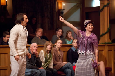 Navarre, in white sweater and pants, reacts to Rosaline, in purple shaw and flapper hat and dress, revealing her love token, with members of the audience watching behind