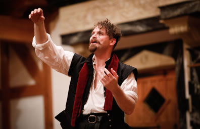 Hamlet looking up, motioning with both arms, wearing white shirt, black renaissance vest and a red scarf draped over his shoulder