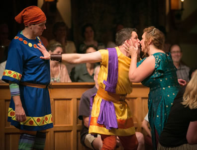 Adriana to right kissing Antipholus, holding his cheeks in her hands, and Antipholus has his arm outstretched behind him with his hand on Dromio's chest, holding him back; Dromio stands there looking puzzled. AUdiences in the background, and the blond wood stage wall behind which audiences sit.