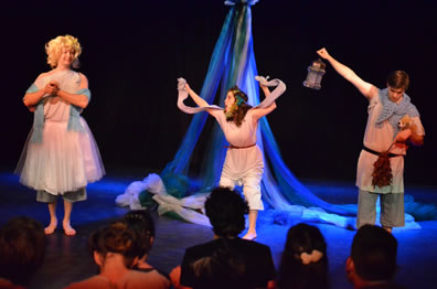 Flute as Thisbe wears a ballerina dress over his shin-length pants and bare feet, a blue scarf draped around his arms, and a blond wig with hands clasped together; Snug as Lion has her hands raised holding the mantle across the back of her neck, flowers in her hair; Starveling as Moonshine holds up the lantern in his right hand, holds a stuffed dog in the crook of his left arm and holding a thorn bush in that hand, his head down. We see the backs of the noble audience's heads. Maypole is in the background.