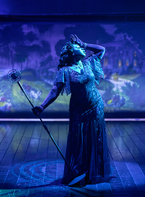 Lady Liv in blue fancy dress and thin cape around shoulders, leans back dramatically as she stands in a blue spotlight  with gloved left hand to her forehead and her outstreched right hand holding the stand of an old microphone. Curier and Ives lithograph of "A Home on the Mississippi" is in the background. Photo by Daniel Rader 