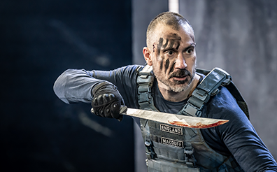 STC-Macbeth-Macduff-3170 Macduff in battle uniform and black charcoal smeared across his face stands with daggar poised.