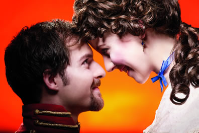 Closeup of Benedick and Beatrice smiling at each other with foreheads touching against a bright orange background.